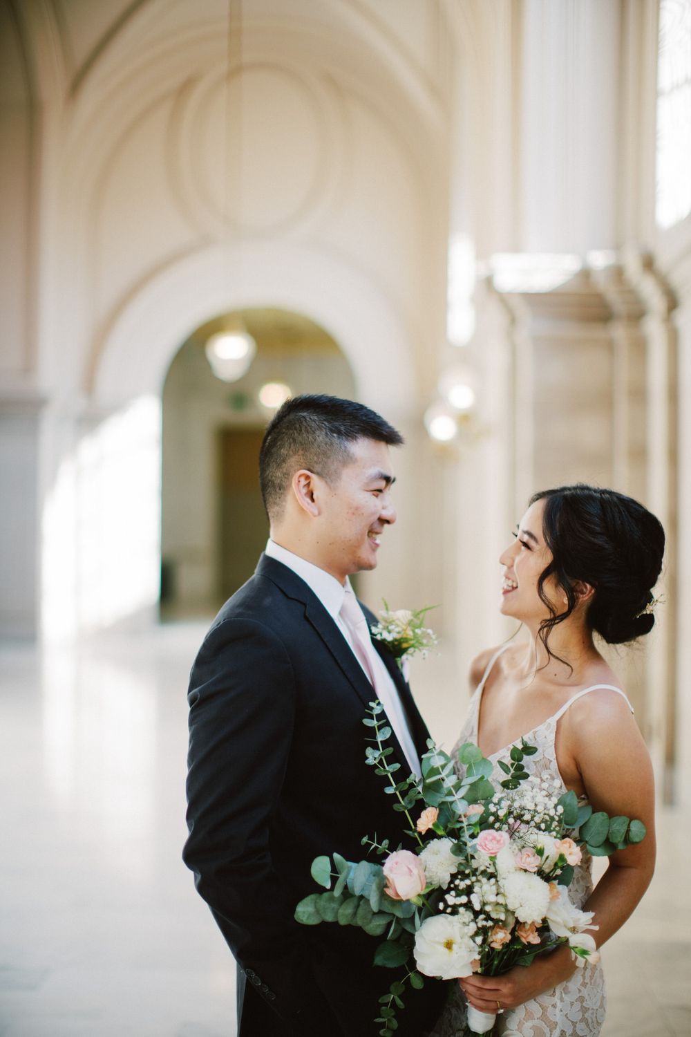 A joyful wedding couple laughs while holding a beautiful bouquet in an elegant hallway.