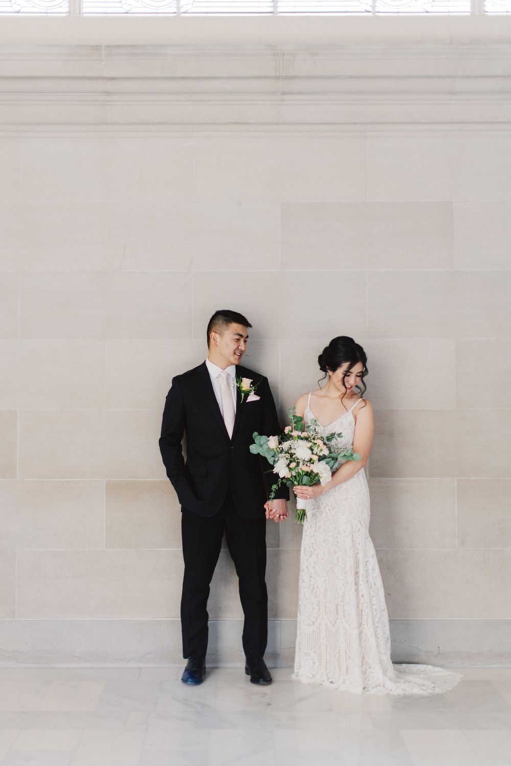 Elegant bride in lace gown and groom in black suit pose against light wall with white floral bouquet.