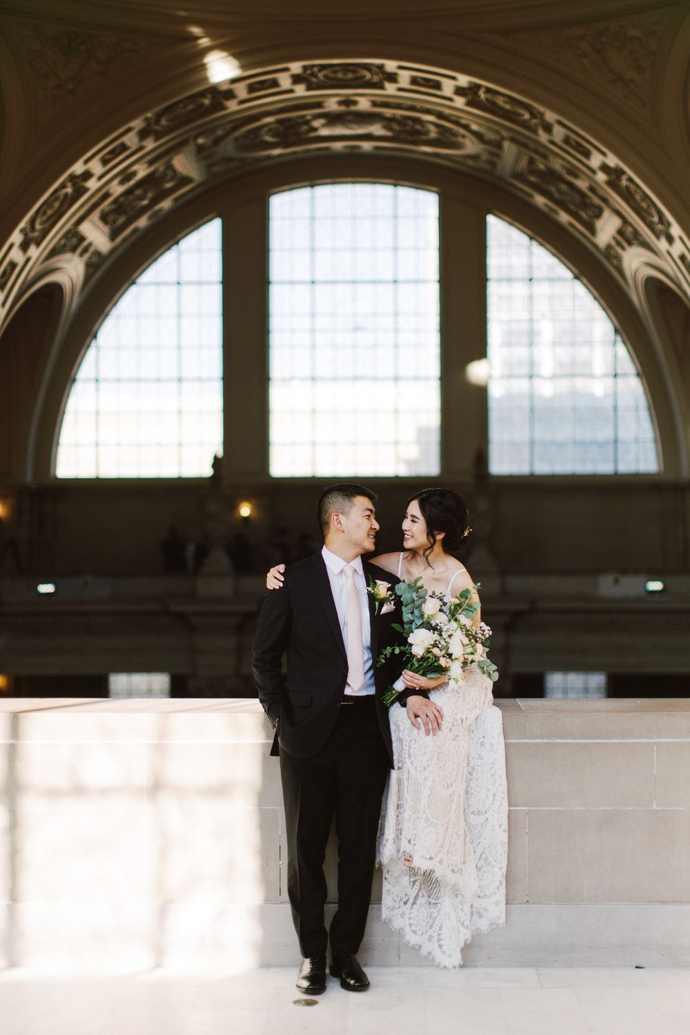 Wedding couple stands beneath ornate archway in historic building with natural light streaming through.