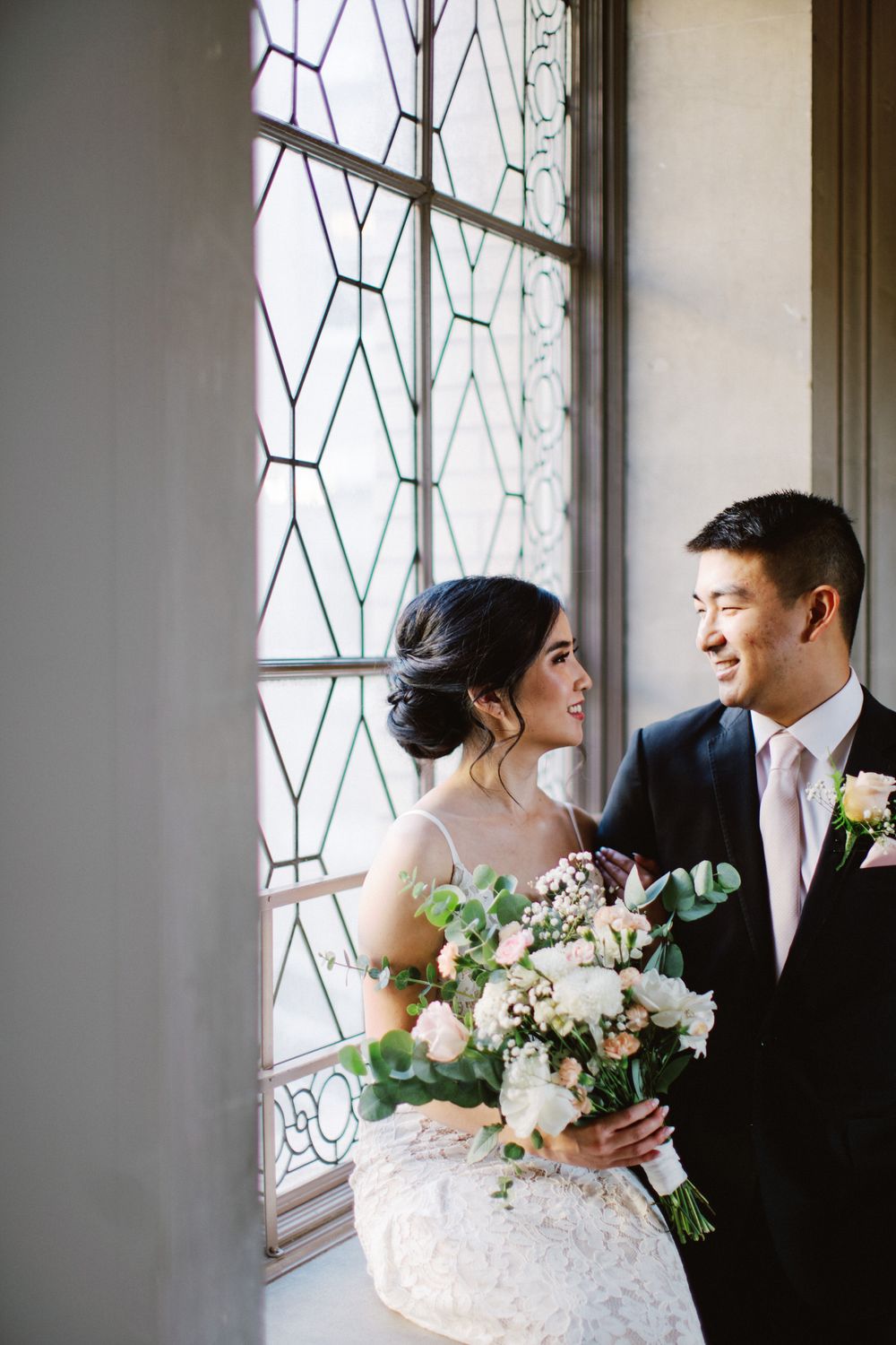 Bride and groom share loving glance by geometric window with stunning bouquet of white flowers.