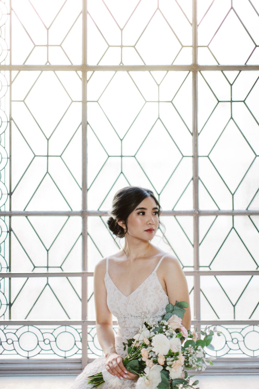 Elegant bride in white lace gown poses by geometric window with delicate floral bouquet.