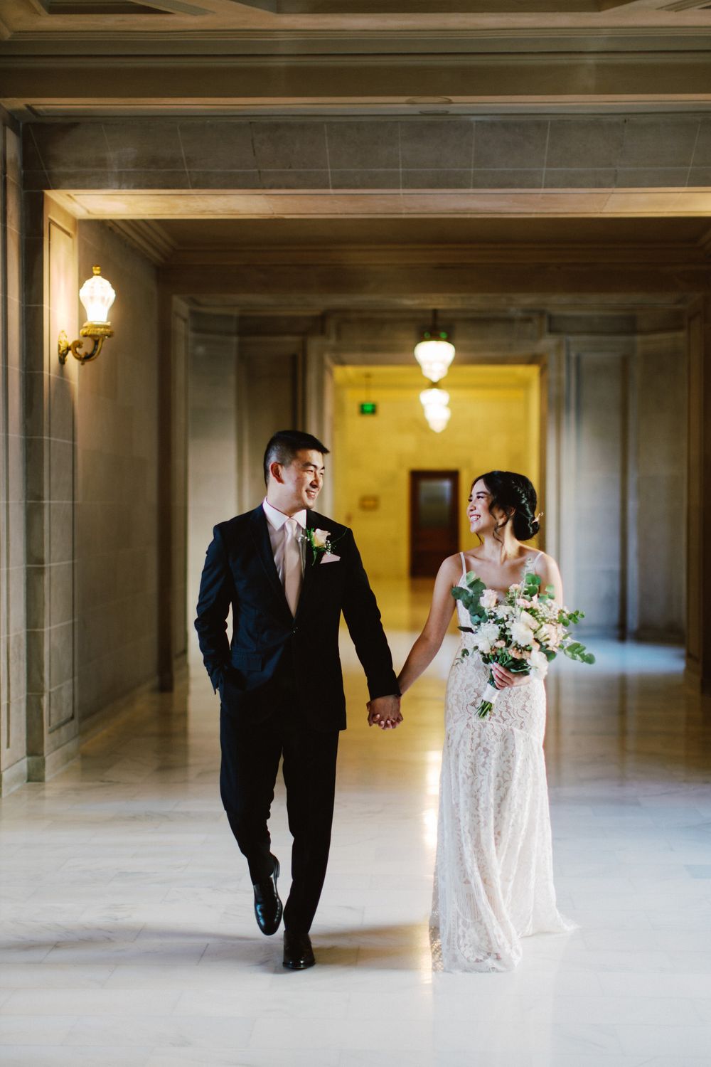 Newlyweds walk hand in hand down elegant hallway with vintage lighting fixtures.