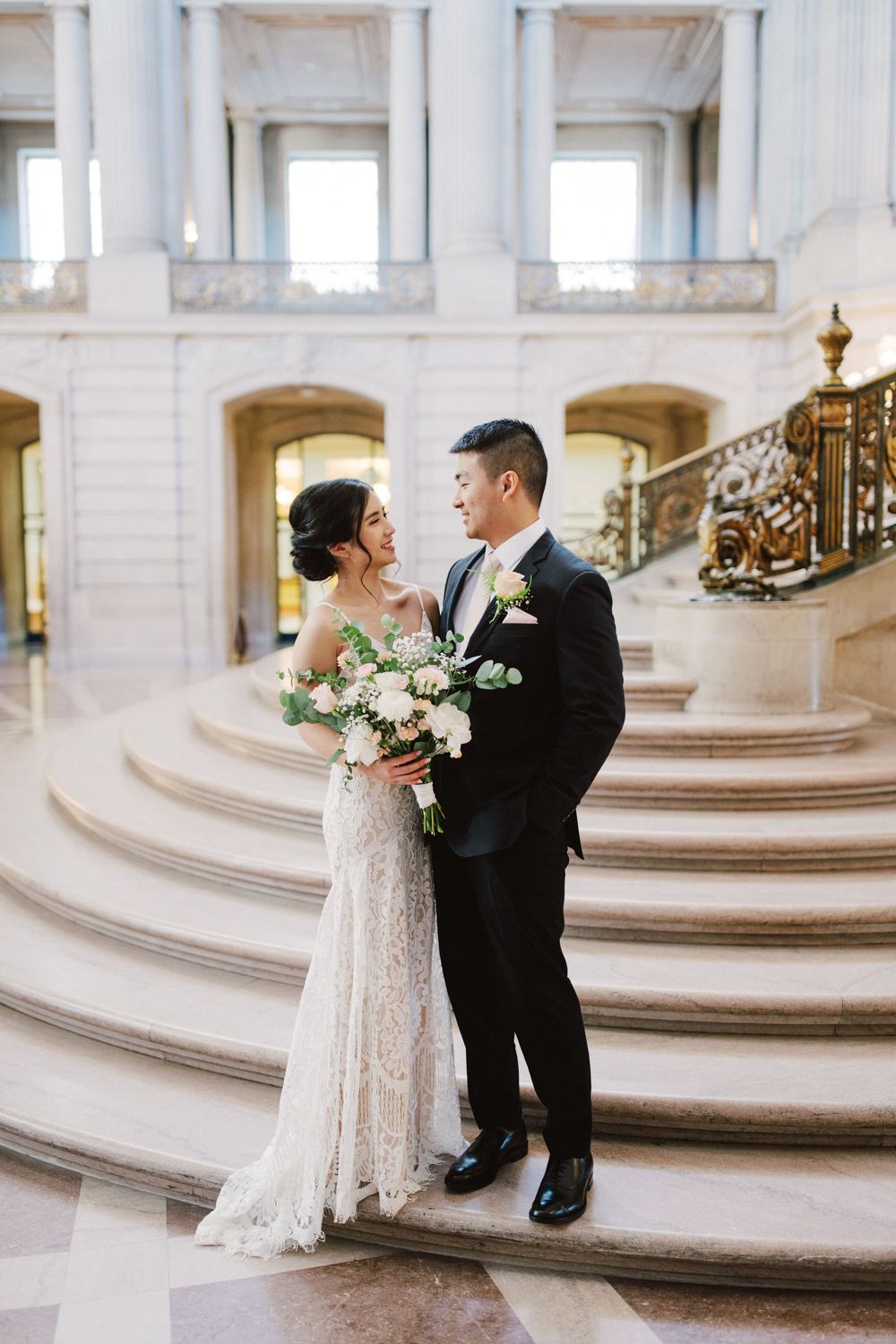 Wedding couple poses on grand marble staircase with ornate brass railing.