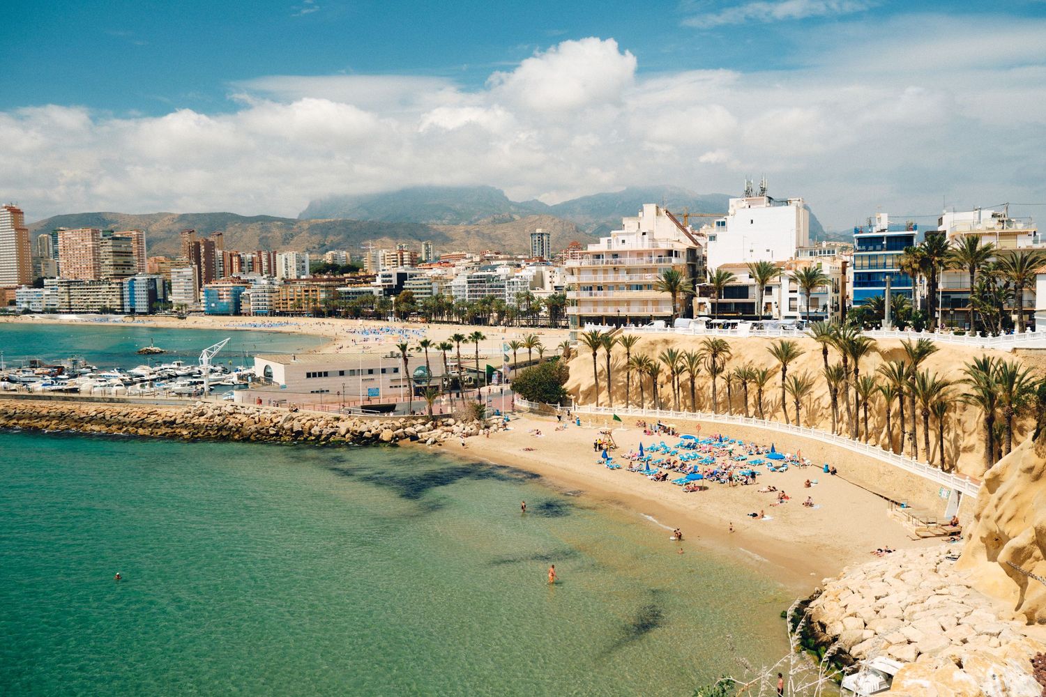 Scenic Mediterranean beach cove with turquoise waters and urban skyline against mountain backdrop.