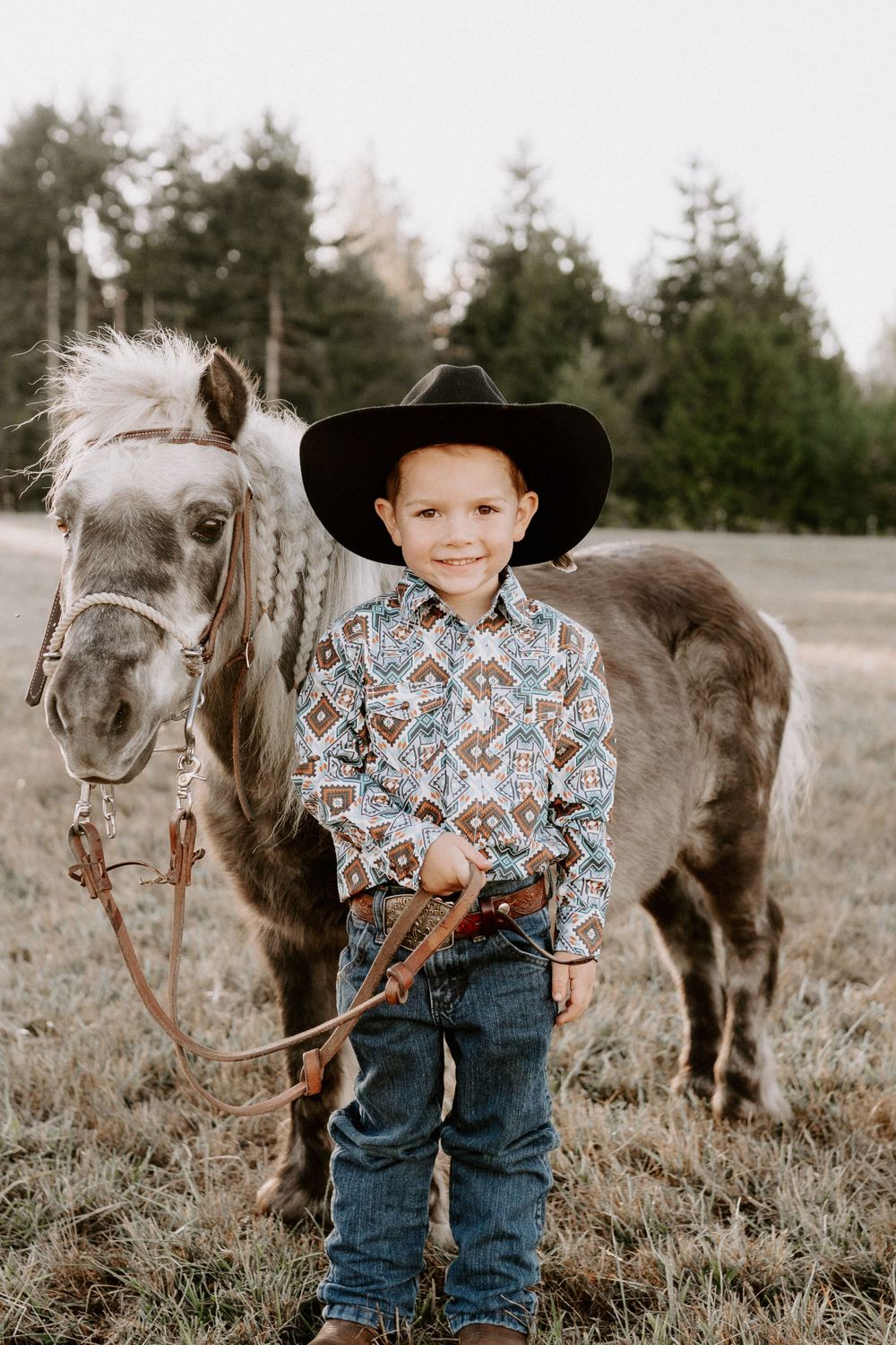 Young boy in cowboy hat and western shirt standing next to gray pony in field.