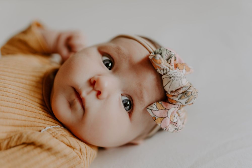 Baby wearing a floral headband and peach colored sweater laying on white bedding.