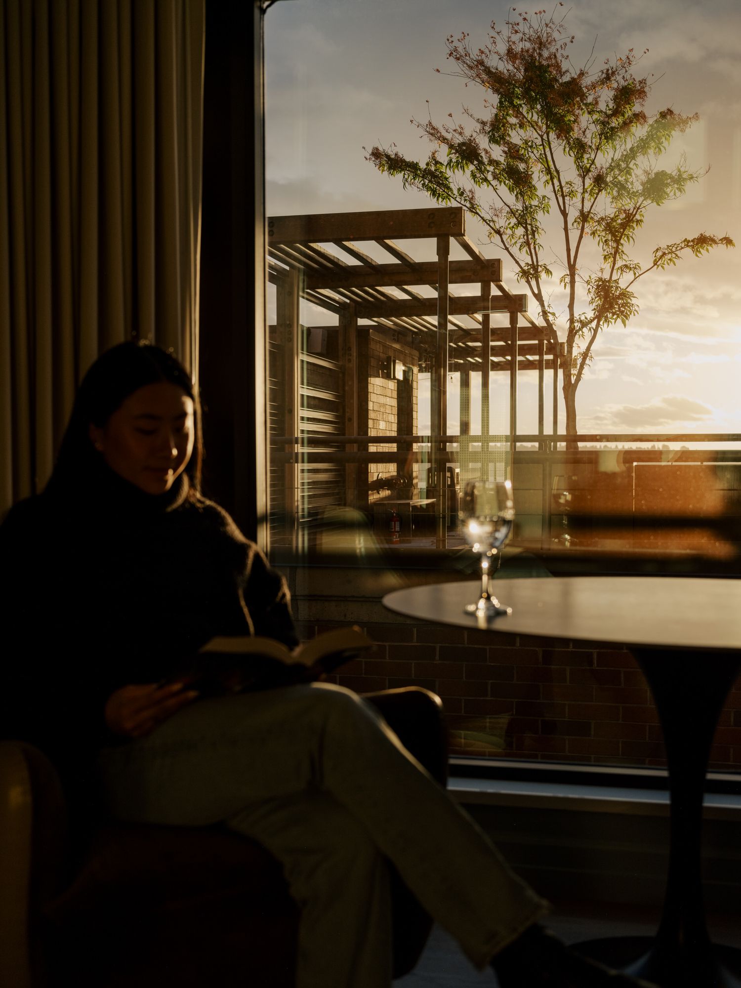 A silhouetted figure sits by a table near a pergola during golden hour sunset, with a wine glass on the table.