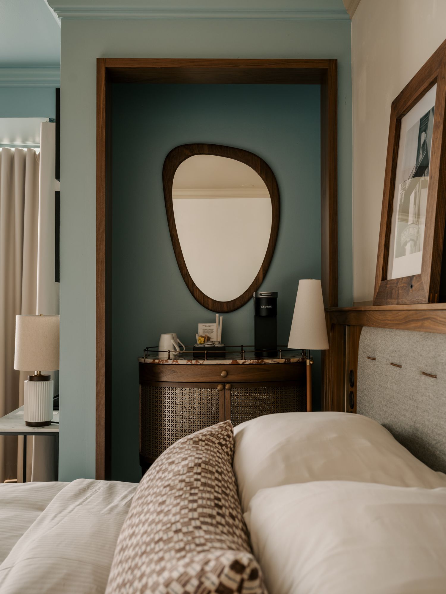 Cozy bedroom interior with teal walls, organic-shaped mirror, and white bedding with knit throw blanket.