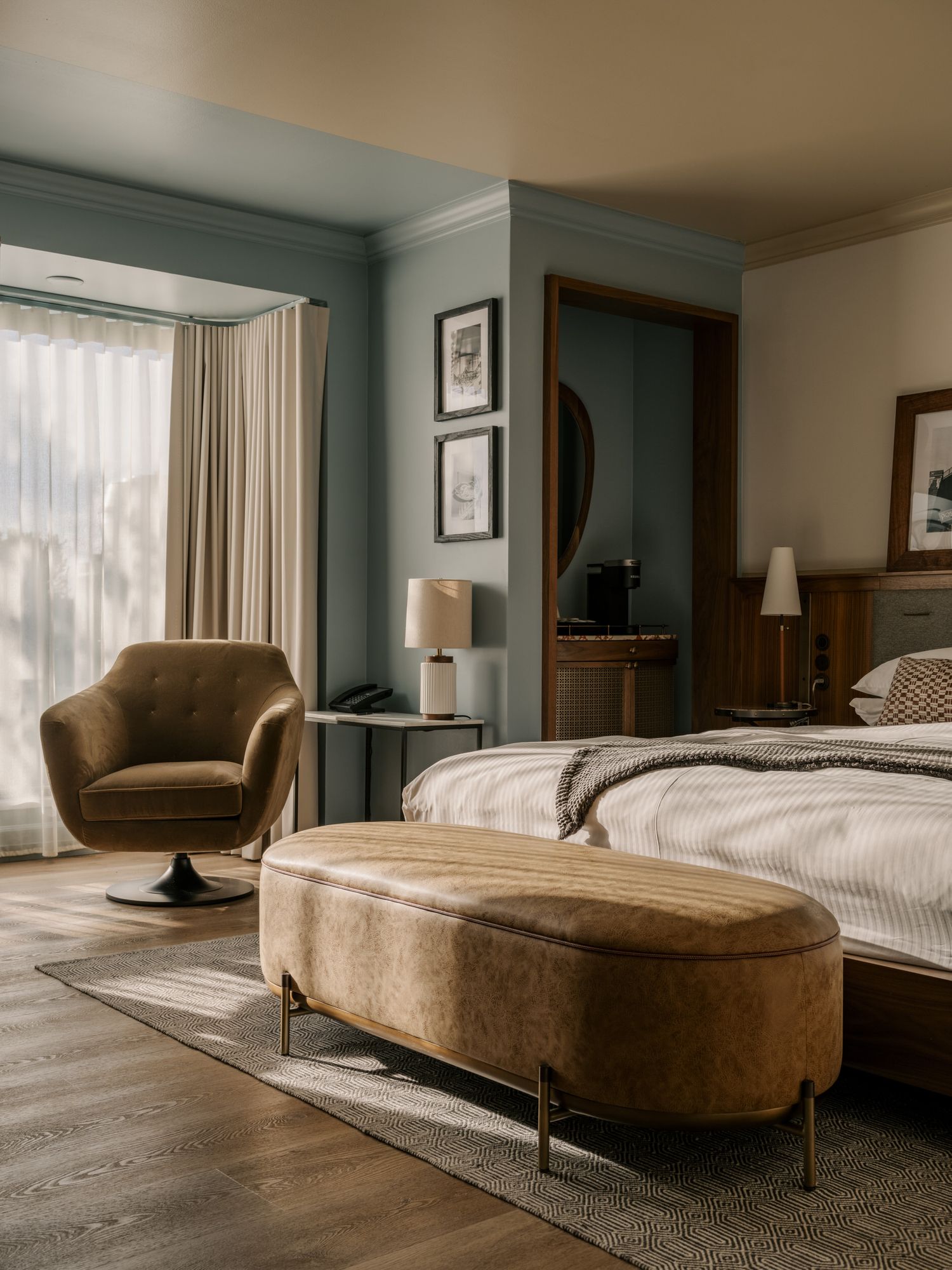 A modern hotel bedroom interior with brown swivel chair, oval ottoman, and soft natural lighting through sheer curtains.