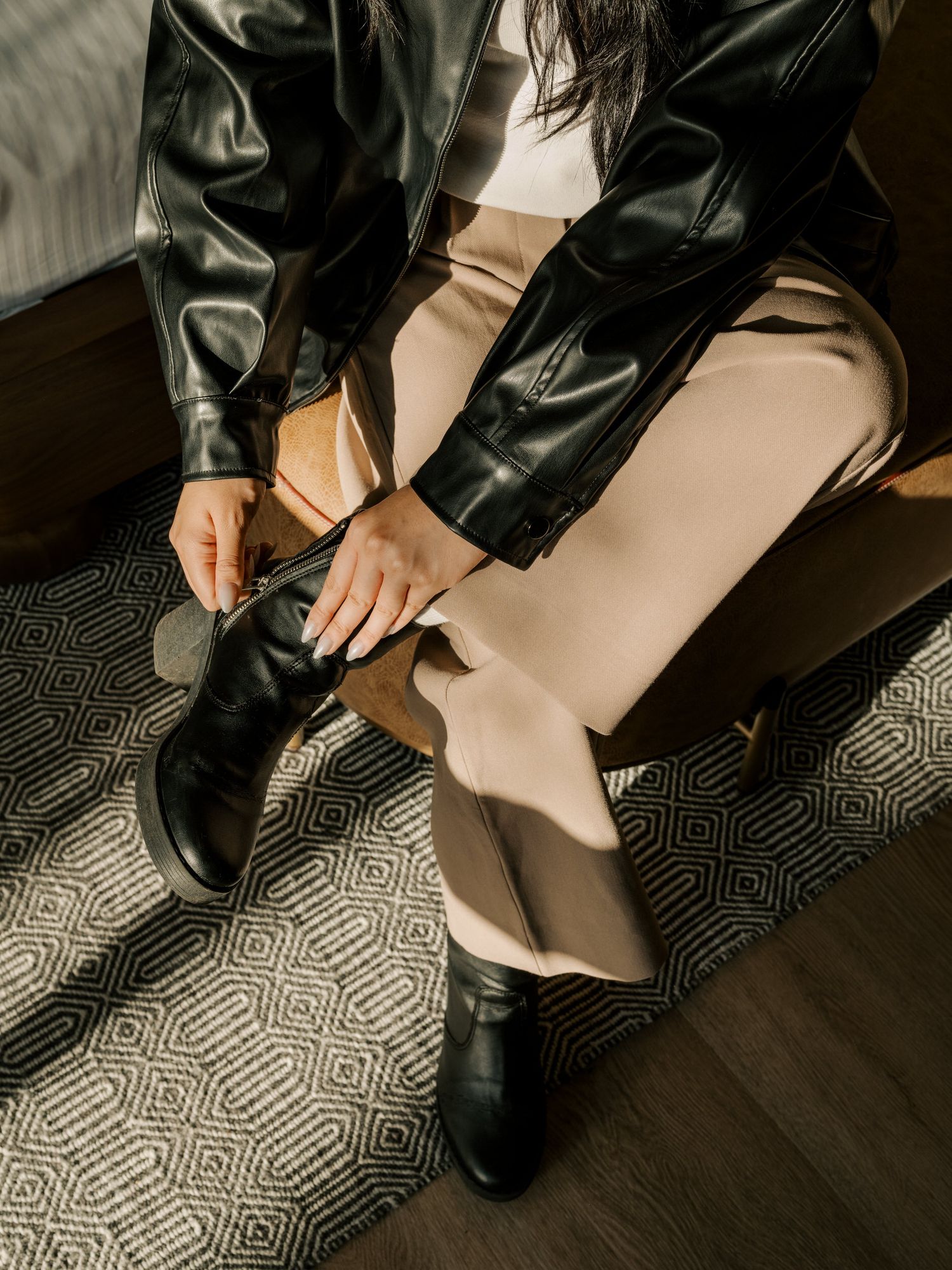 Someone tying black leather boots while wearing a leather jacket and beige pants on a patterned floor.