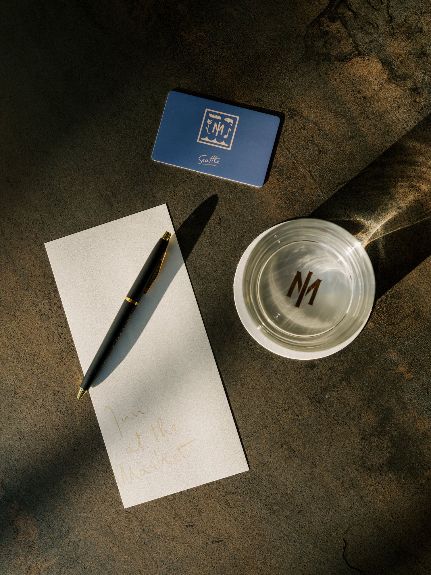 A minimalist workspace with a glass of water, blank notepad, black pen, and blue business card on a textured surface.