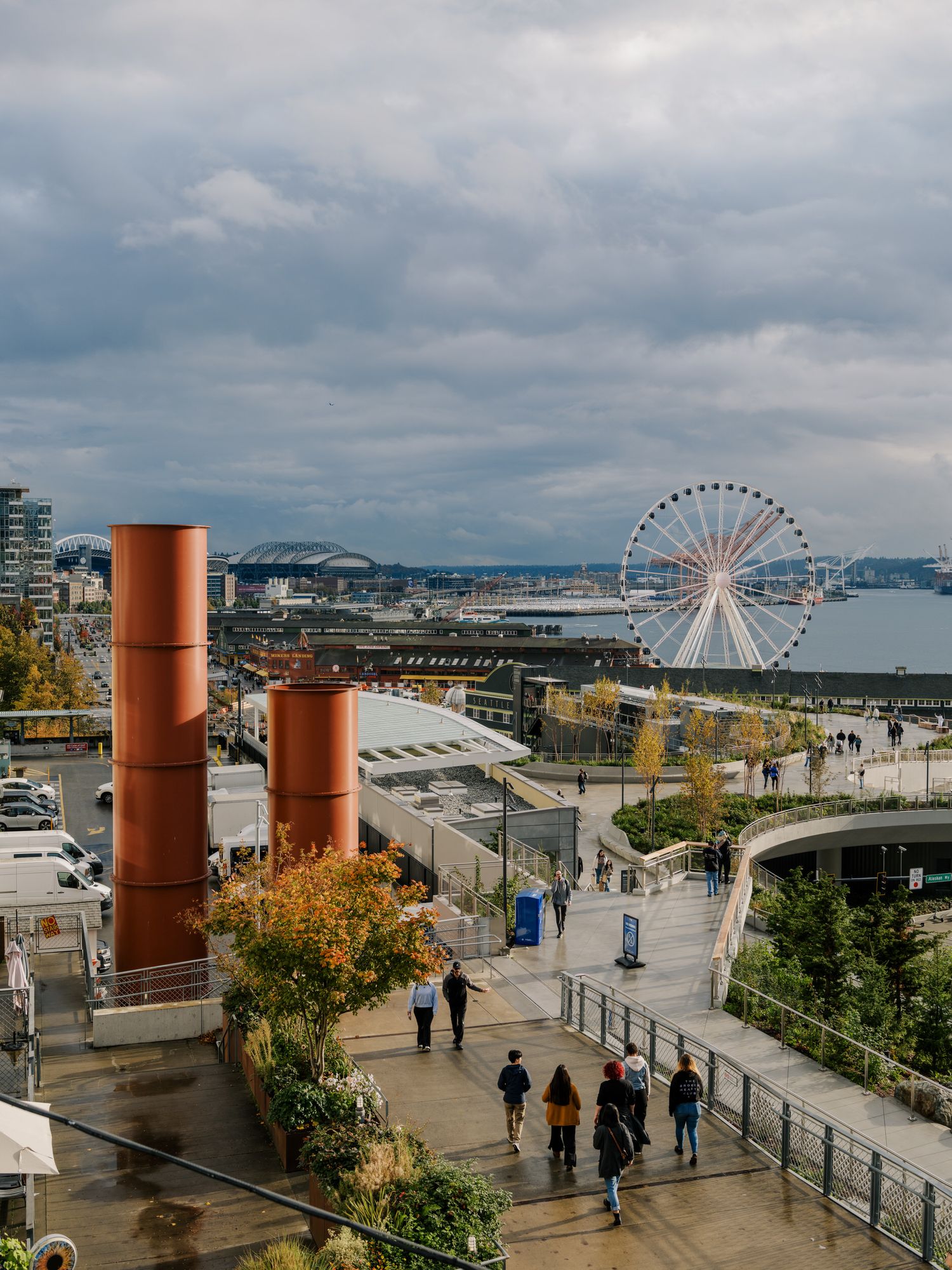 Orange industrial smokestacks stand near Seattle's waterfront with the Great Wheel visible against a cloudy sky.