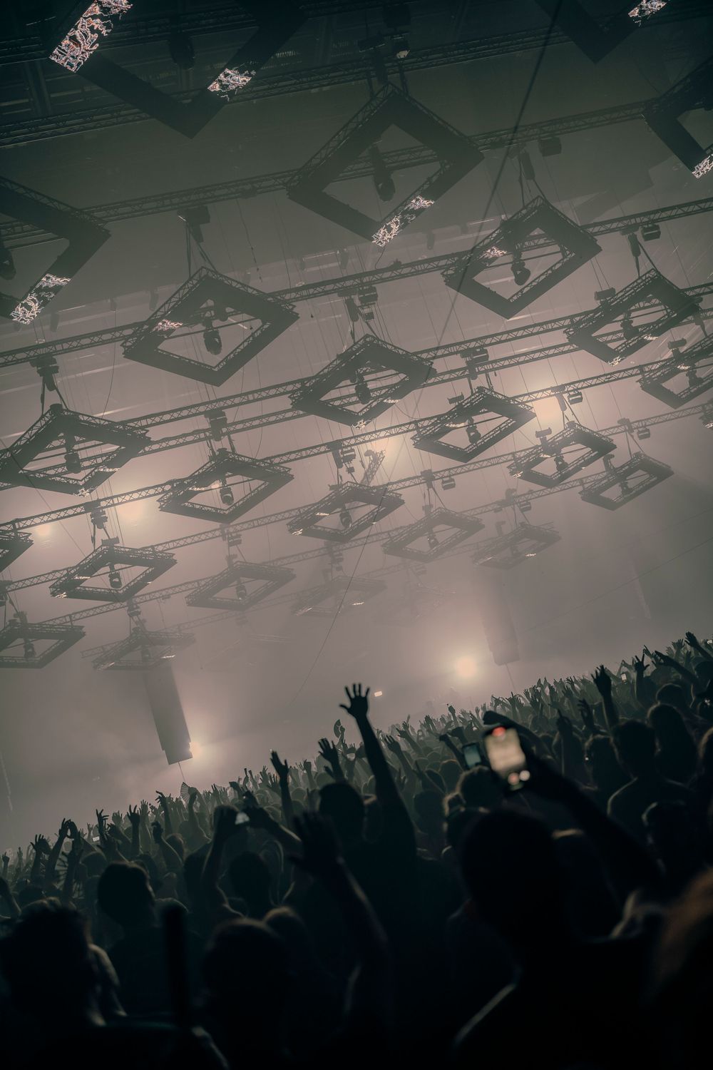 Silhouetted crowd with raised hands at a packed concert venue with dramatic overhead lighting.