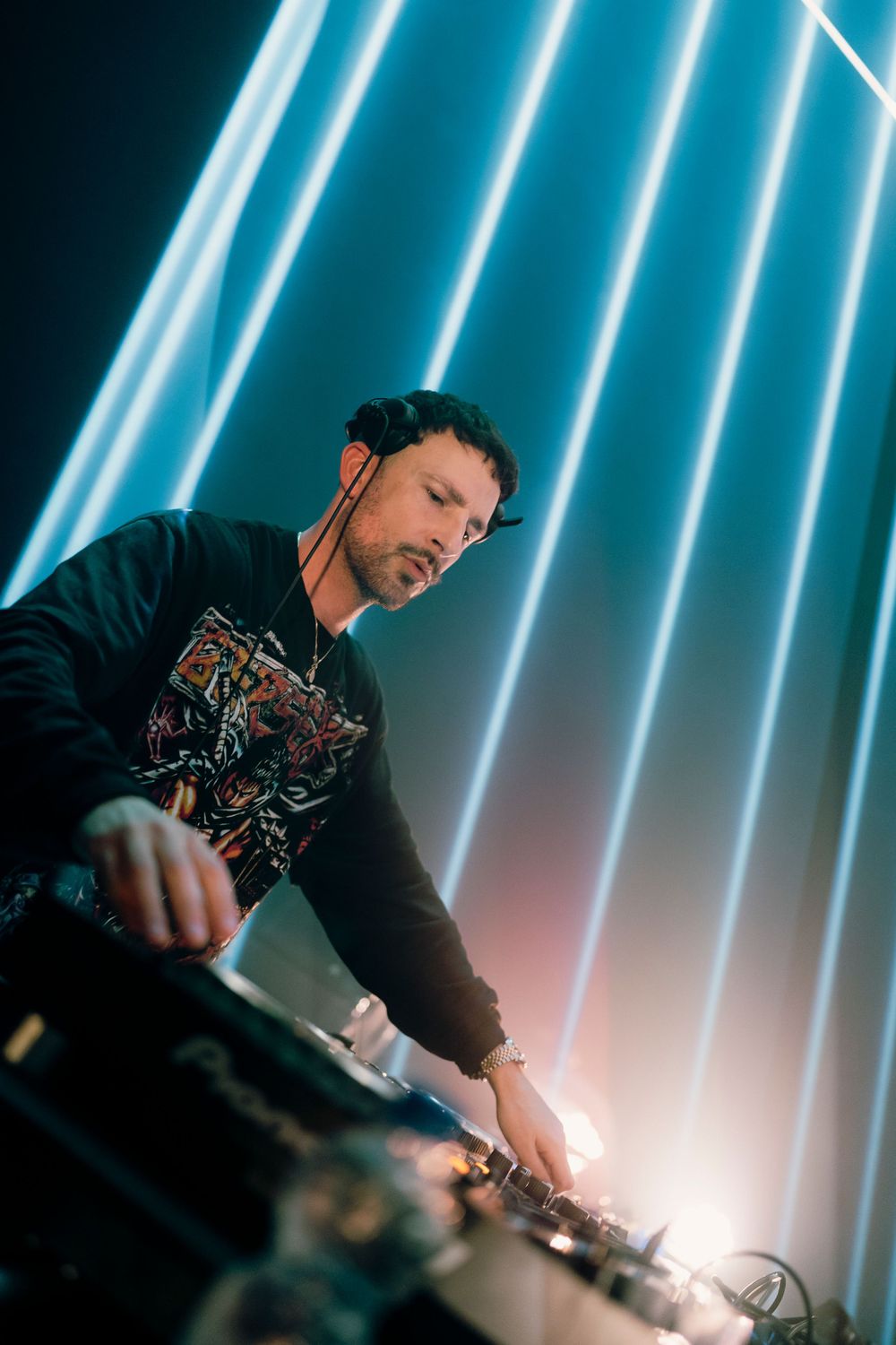 DJ performing behind decks with striking vertical neon blue light beams in background.