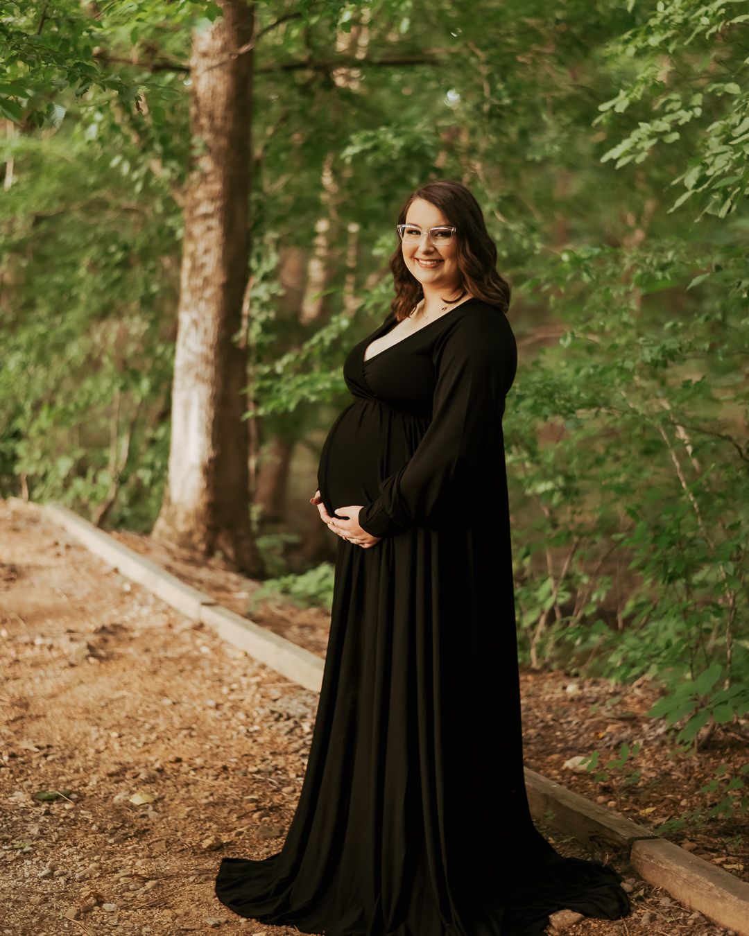 Outdoor maternity session with woman in long black gown on wooded path.