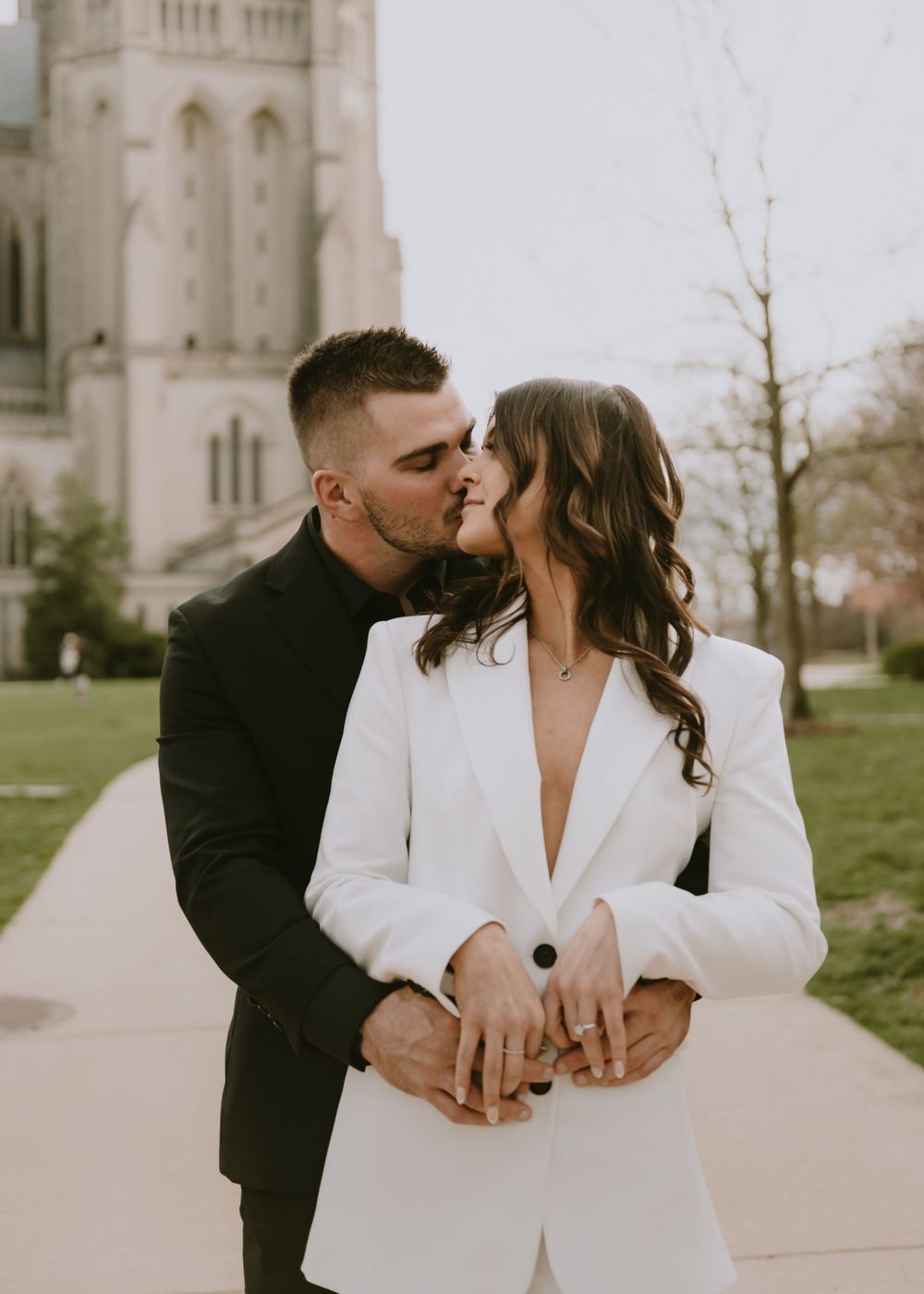 Two people embrace in front of a majestic cathedral, the woman wearing an elegant white blazer.