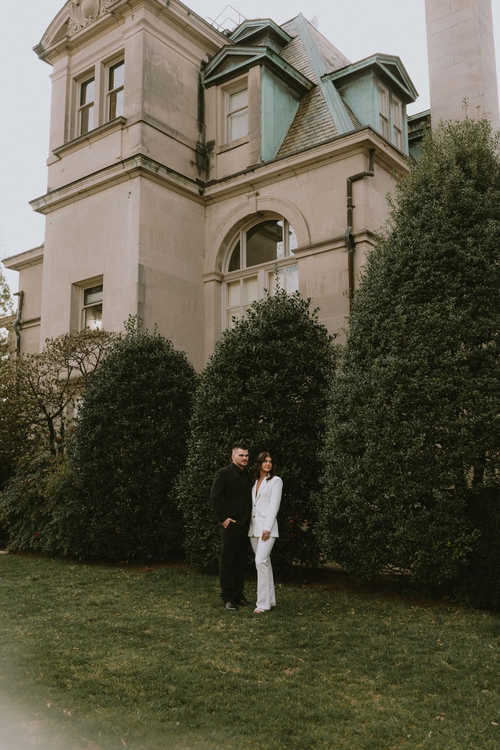A couple poses in formal attire against a historic mansion with manicured shrubs.