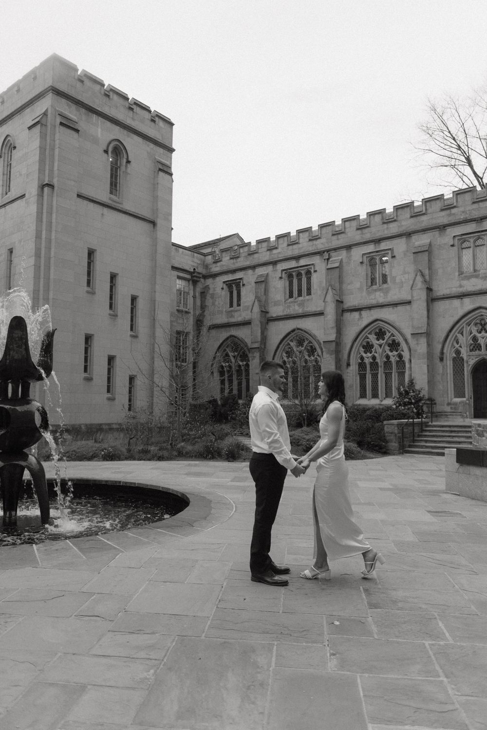 Black and white photo of couple holding hands in courtyard of historic stone church building.