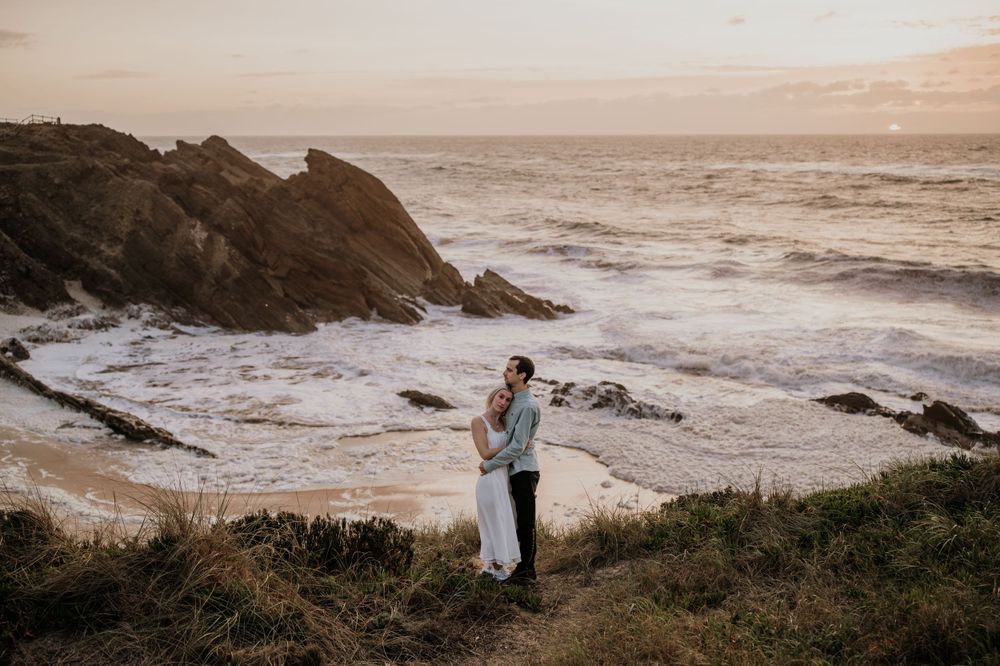 A couple embraces on a dramatic coastal cliff during golden hour with crashing waves below.