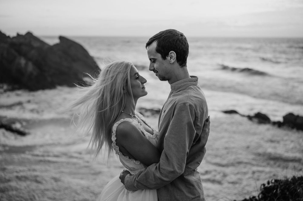 A black and white portrait of a couple in an intimate moment on a windswept coastal cliff.