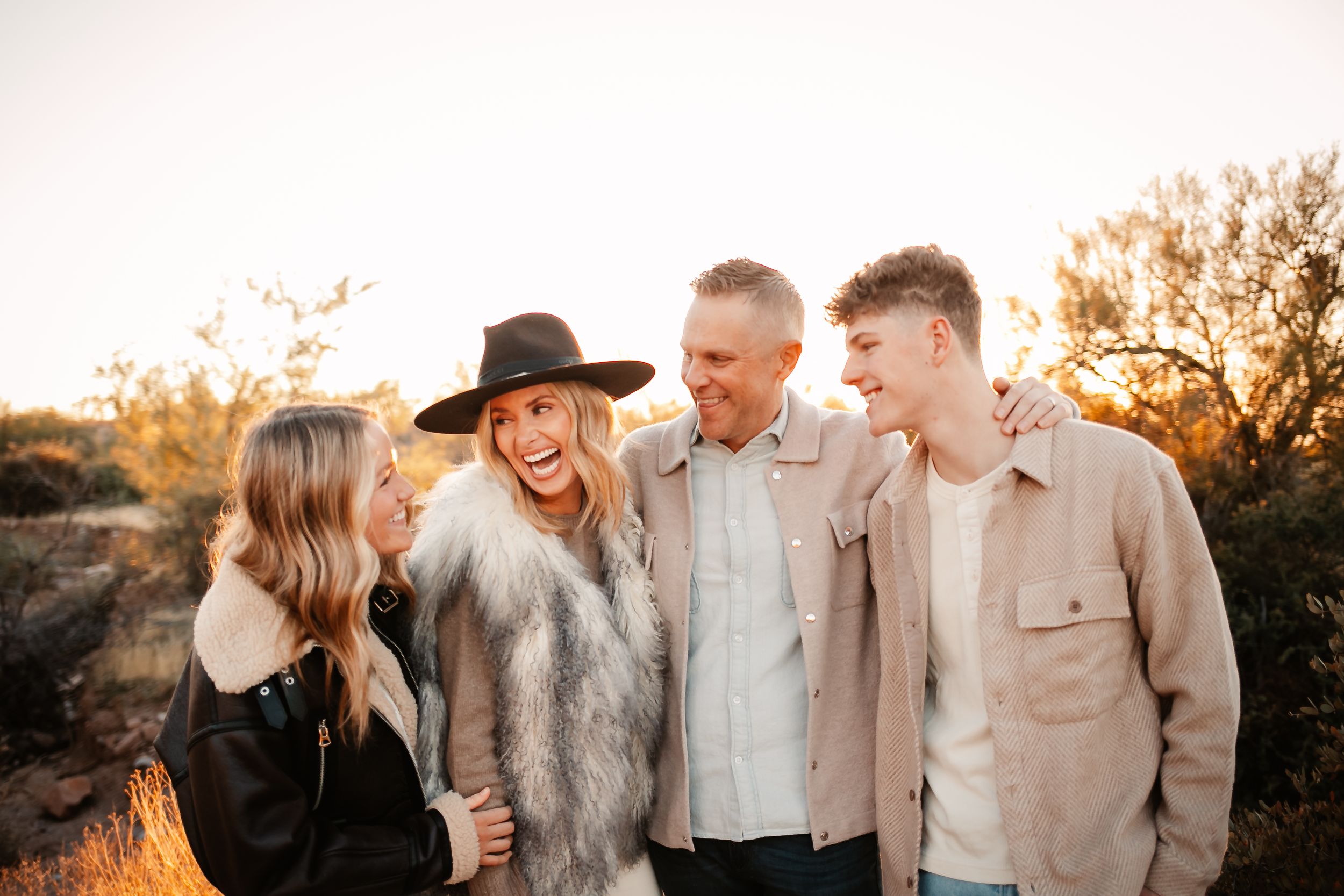 A laughing family of four embraces outdoors during a warm golden hour sunset in a desert setting.