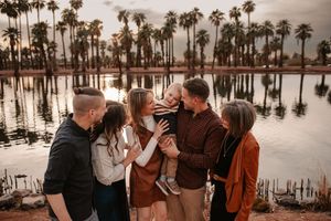 Family of six gathered by a palm tree-lined lake at sunset, sharing a joyful moment with a toddler in warm autumn tones.