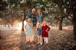 Family of five posing outdoors in an orchard during golden hour, parents with three young children in coordinated fall outfits.