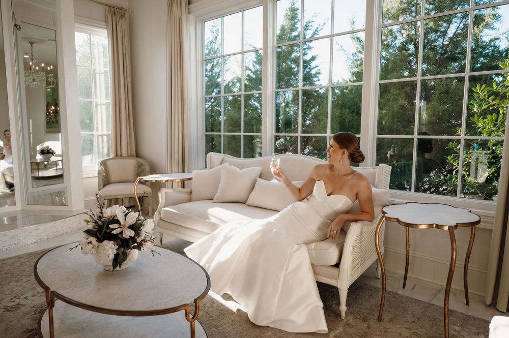 Bright sunlit room with white furniture and large windows overlooking pine trees.