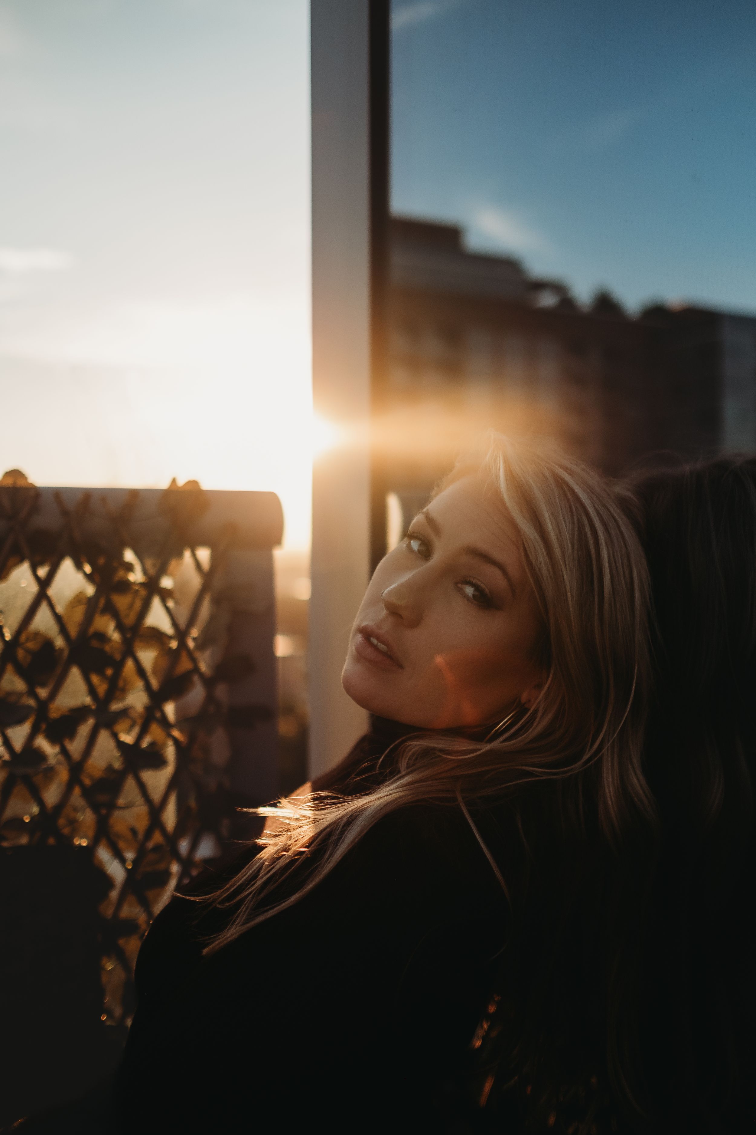 Blonde woman poses near balcony railing bathed in golden sunset lens flare light.