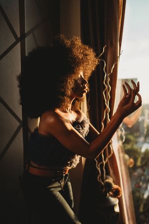 Woman with curly afro gazes out sunlit window while holding sheer curtain in warm light.