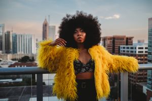 Woman with afro in yellow faux fur coat poses on rooftop with city skyline at dusk.