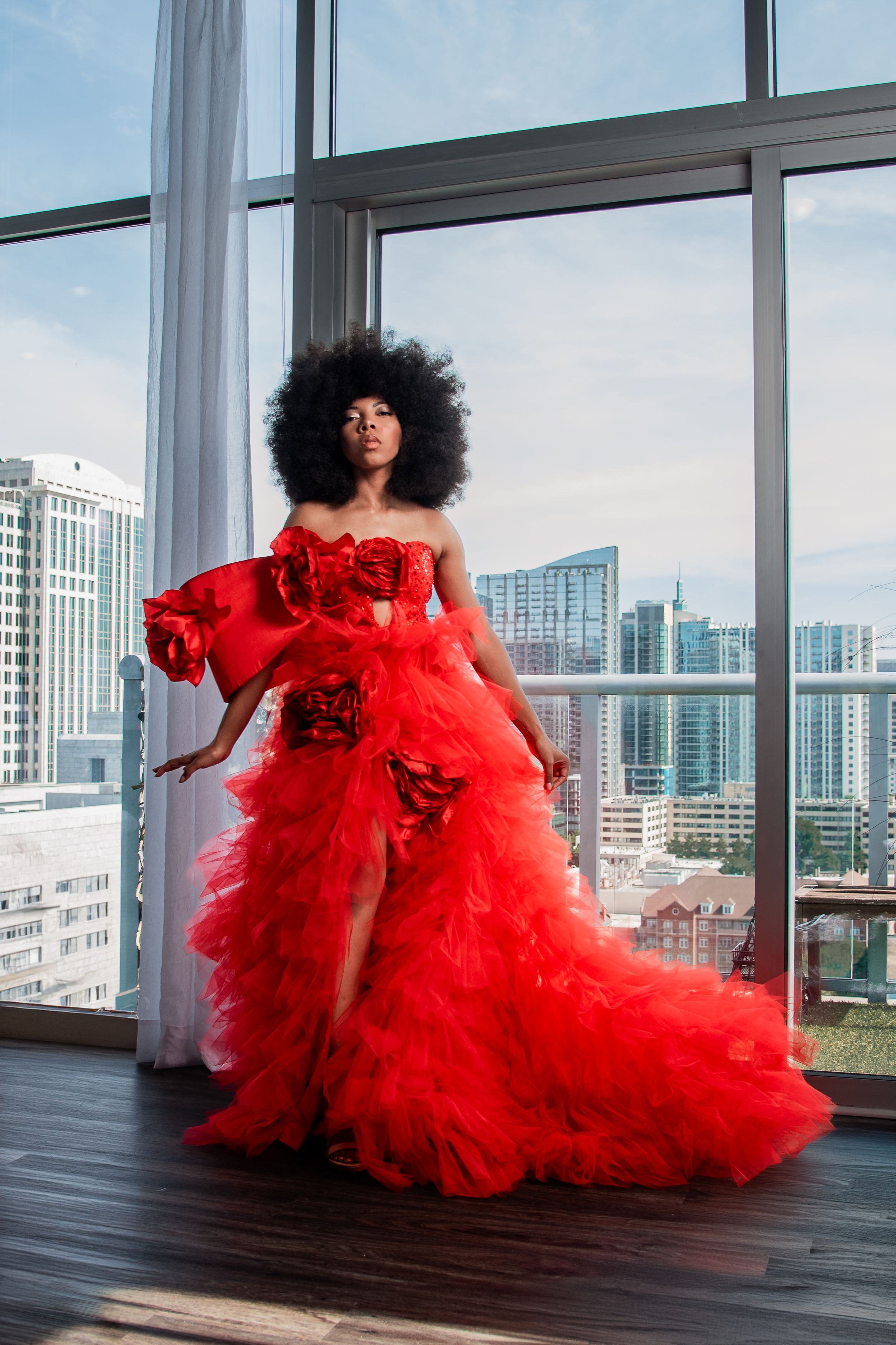Woman in stunning red tulle gown with floral details poses before city skyline window.
