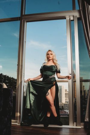 Blonde woman in elegant green velvet off-shoulder gown stands in floor-to-ceiling glass doorway.