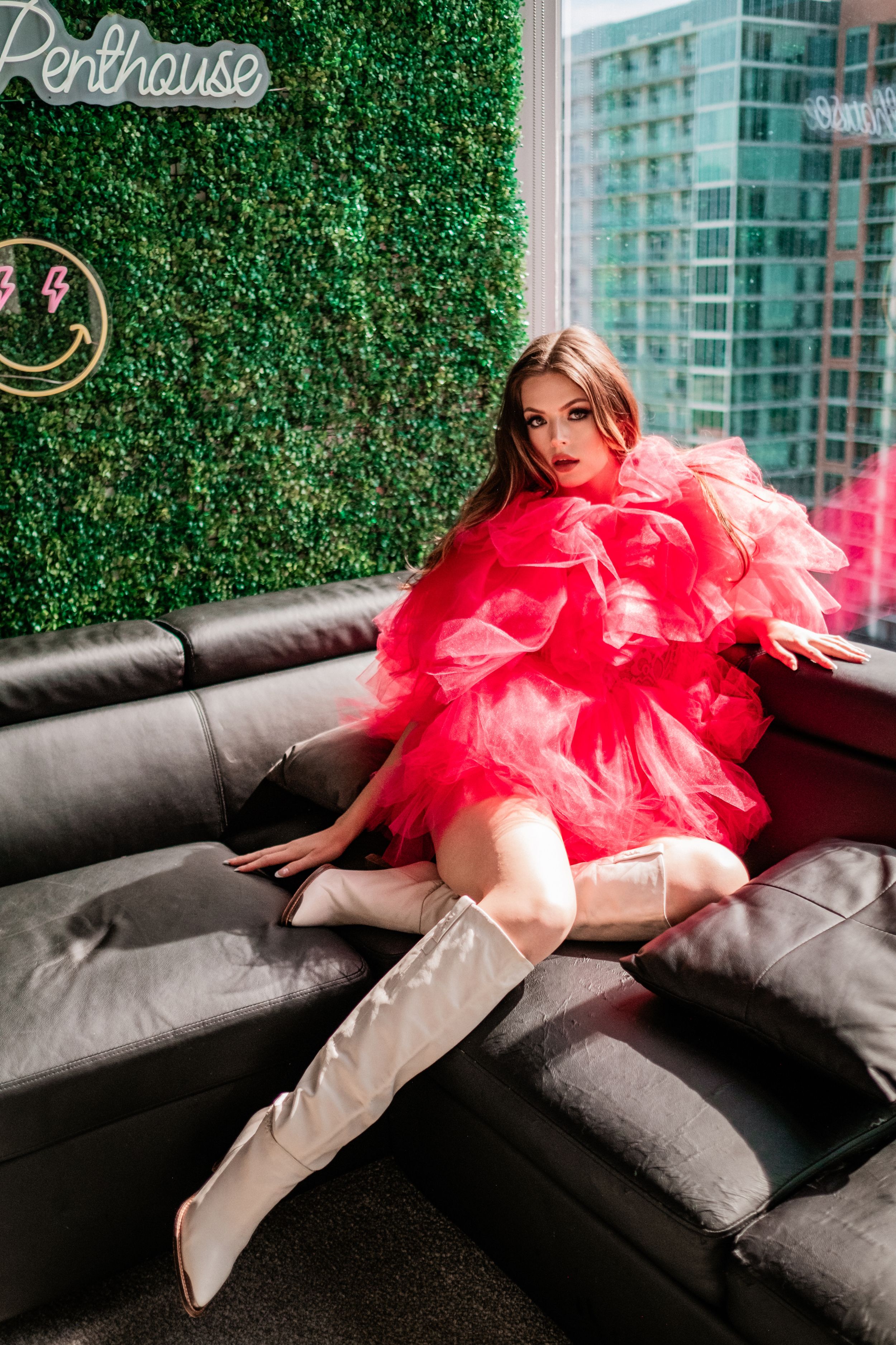 Model in pink ruffled tulle mini dress and white boots poses on black sofa at penthouse.