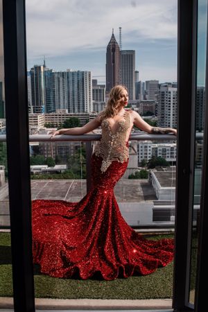 Blonde model in red sequin mermaid gown poses on balcony with Atlanta city skyline backdrop.
