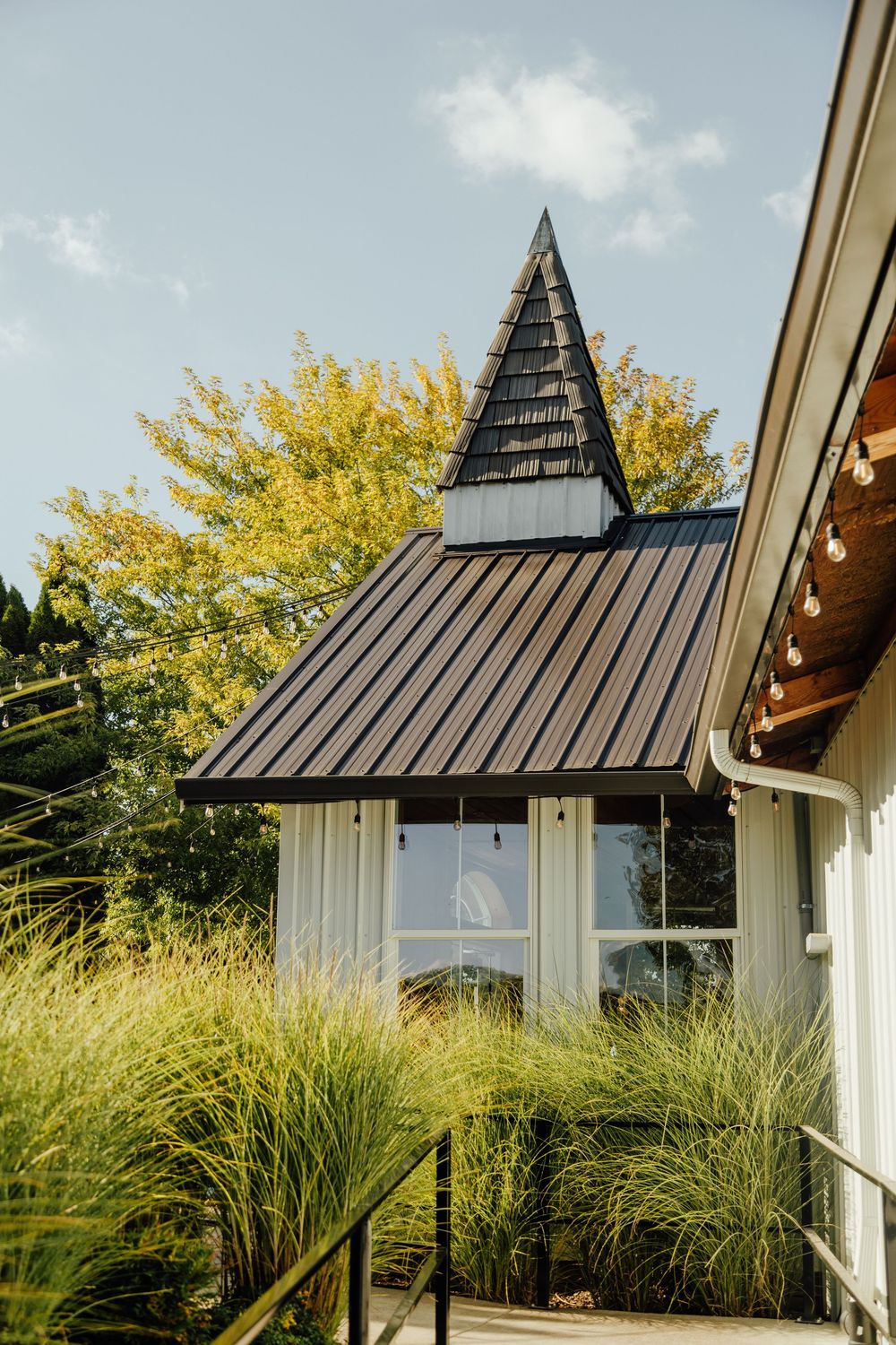 Modern white farmhouse with black metal dormers and architectural details framed by autumn trees and ornamental grasses.