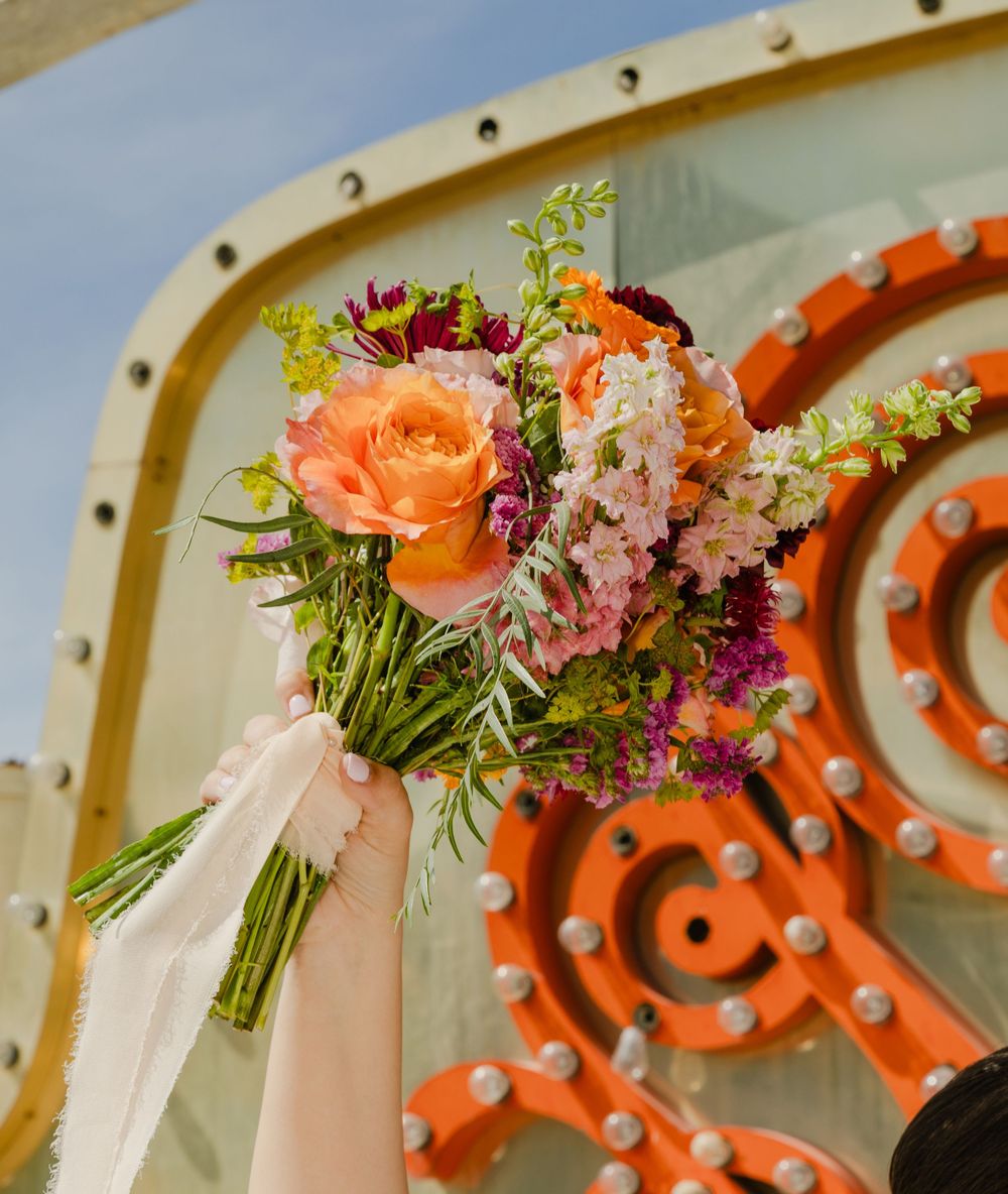 Colorful bridal bouquet with orange roses and wildflowers held against an orange industrial backdrop.