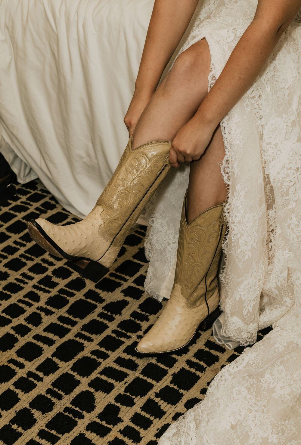 Bride wearing ivory cowboy boots with floral embroidery under a lace wedding dress, sitting on a bed with a patterned rug.