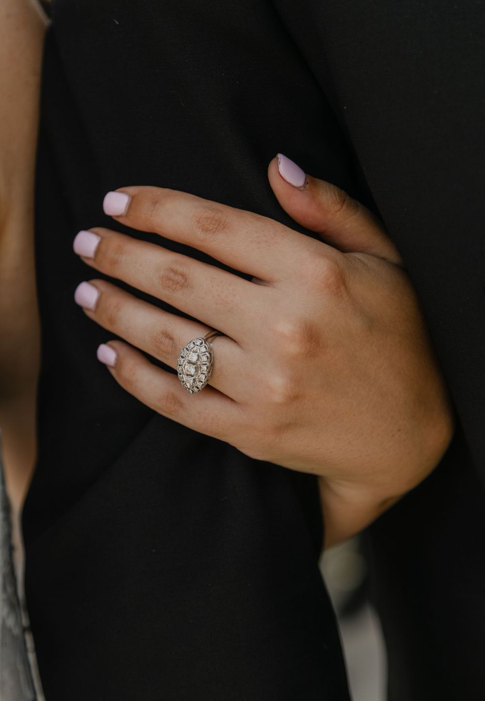 Close-up of a woman's hand with pink nails wearing an elegant diamond cluster engagement ring on her ring finger.