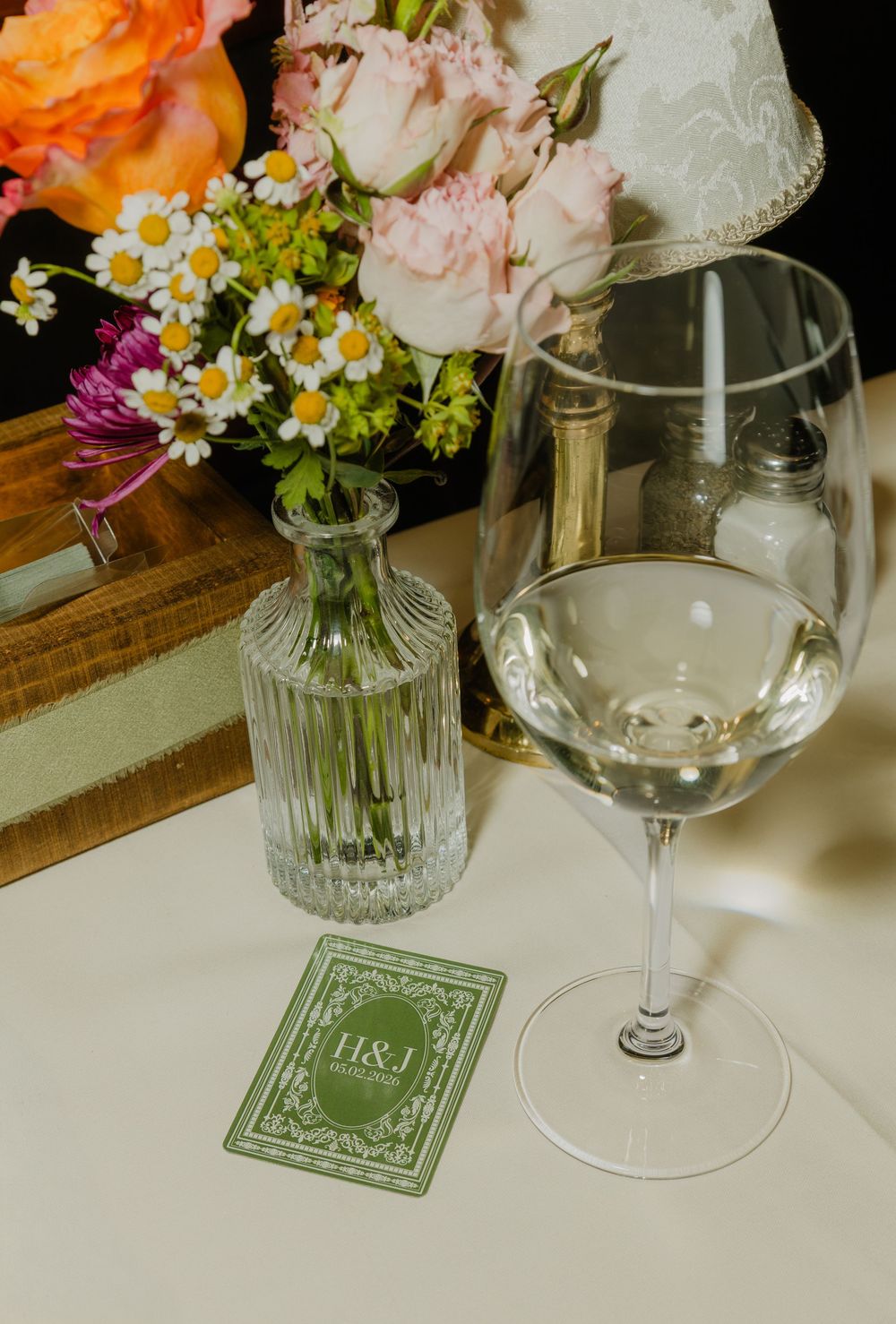 Elegant wedding table setting with wine glass, H&J monogram card, floral arrangement, and glass vase with daisies.