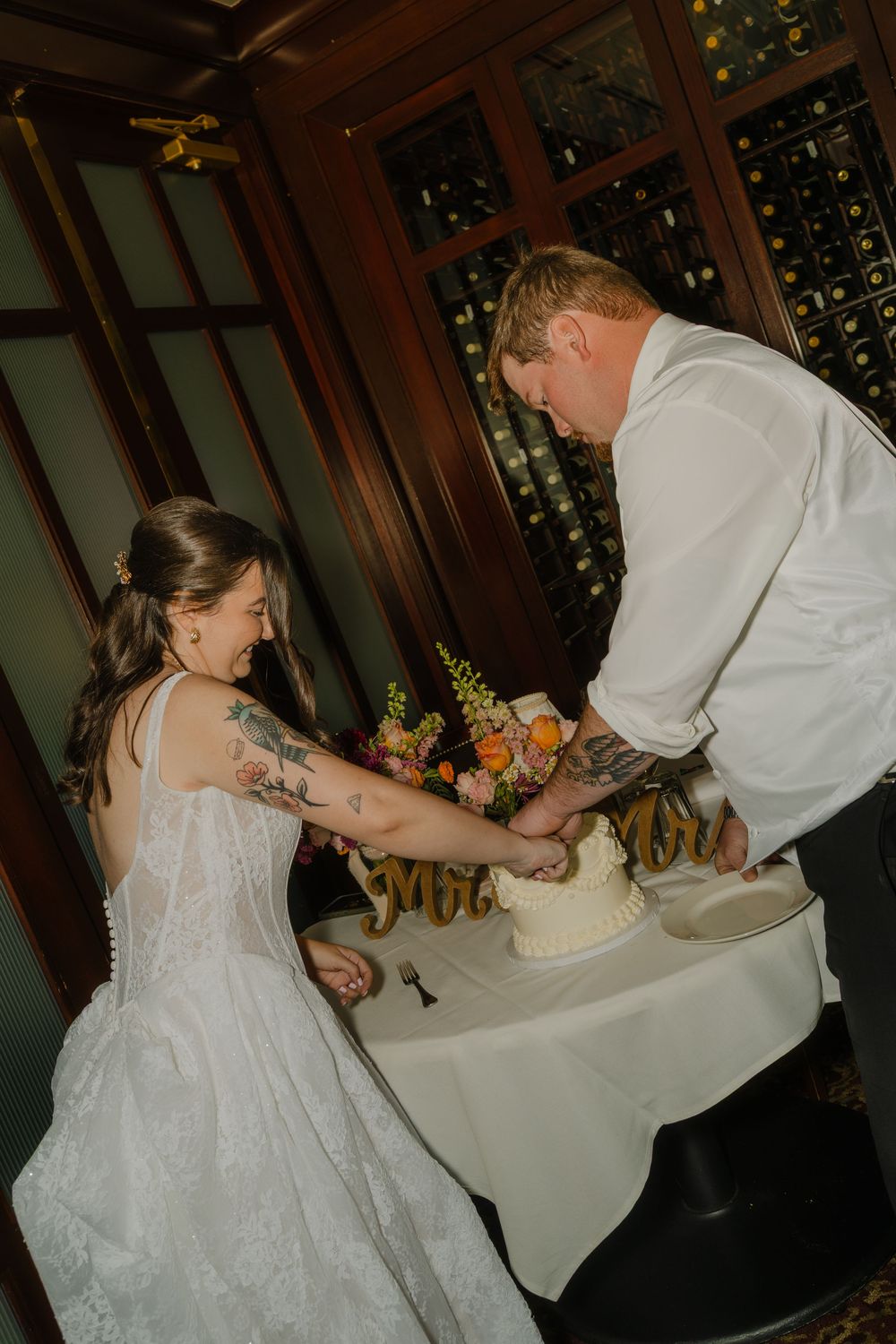 Bride and groom cutting their white wedding cake together in an elegant wood-paneled venue with wine storage.