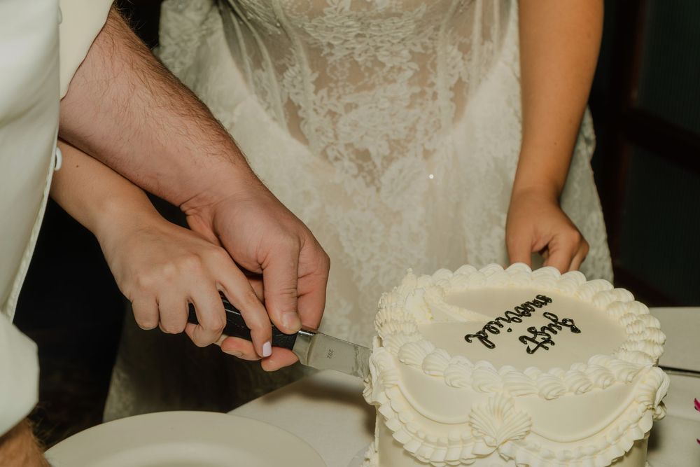 Bride and groom cutting a white wedding cake together, with elegant lace dress visible in the background.