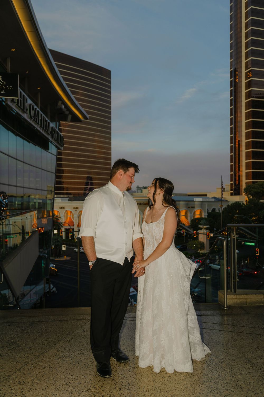 A couple in wedding attire holds hands on a rooftop terrace at dusk with city buildings glowing behind them.