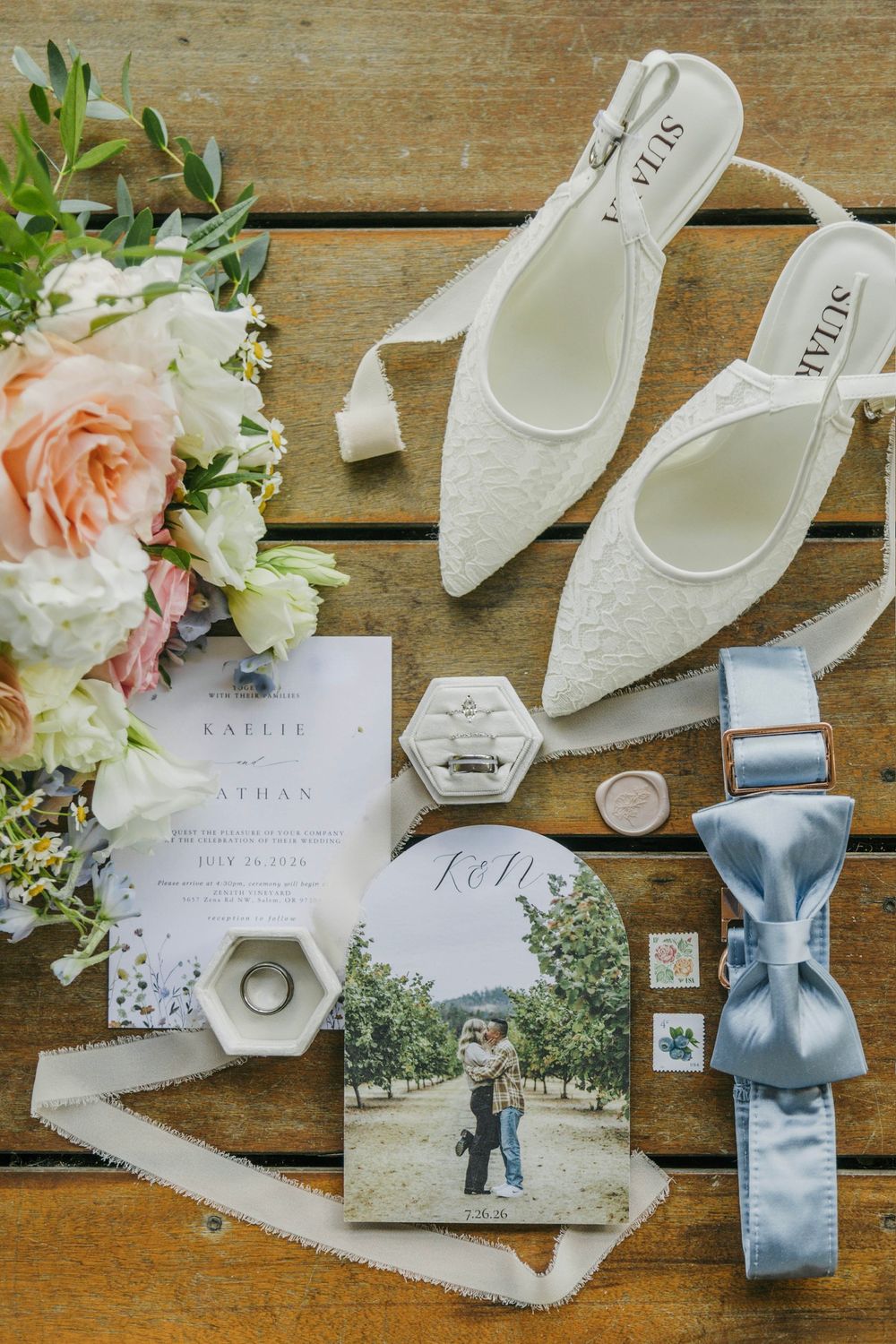 Elegant wedding flat lay featuring white lace heels, floral bouquet, invitations, rings, and a blue bow tie on wood.