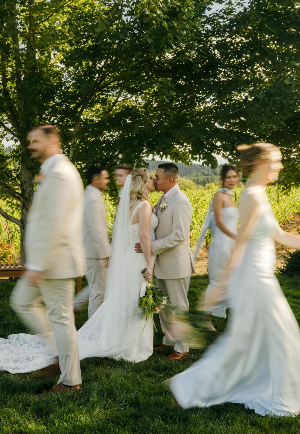 A bride and groom share a romantic moment surrounded by their wedding party in cream attire outdoors.