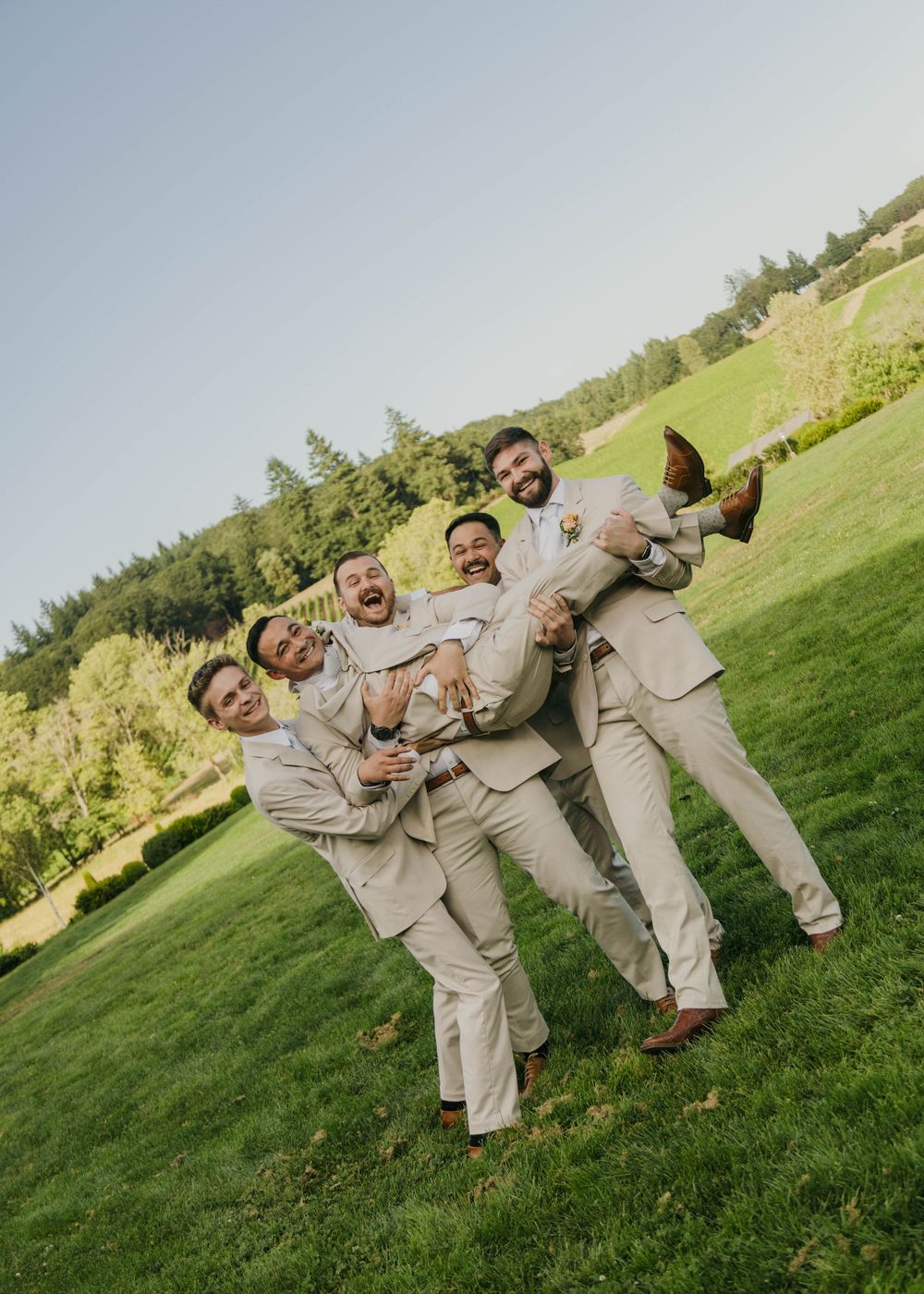 Groomsmen in tan suits joyfully lifting the groom outdoors on a green lawn at a sunny wedding celebration.