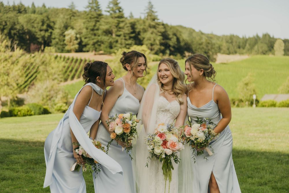 Bride and three bridesmaids in light blue gowns laughing together outdoors at a vineyard wedding.