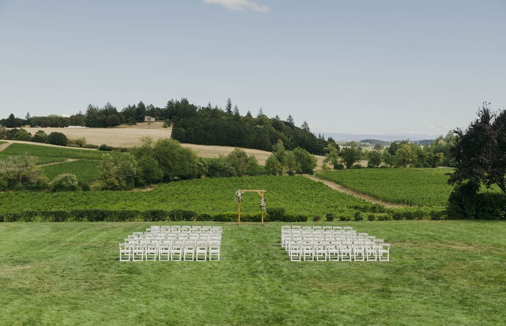 Outdoor wedding ceremony setup with white chairs on green lawn overlooking scenic vineyard and rolling hills.