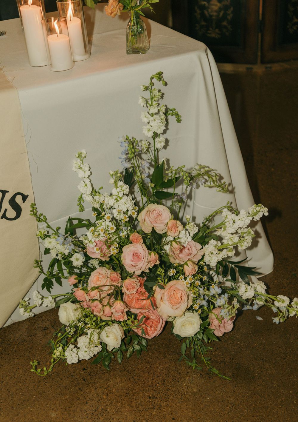Elegant wedding floral arrangement with pink roses and white blooms beside candles on a white tablecloth.