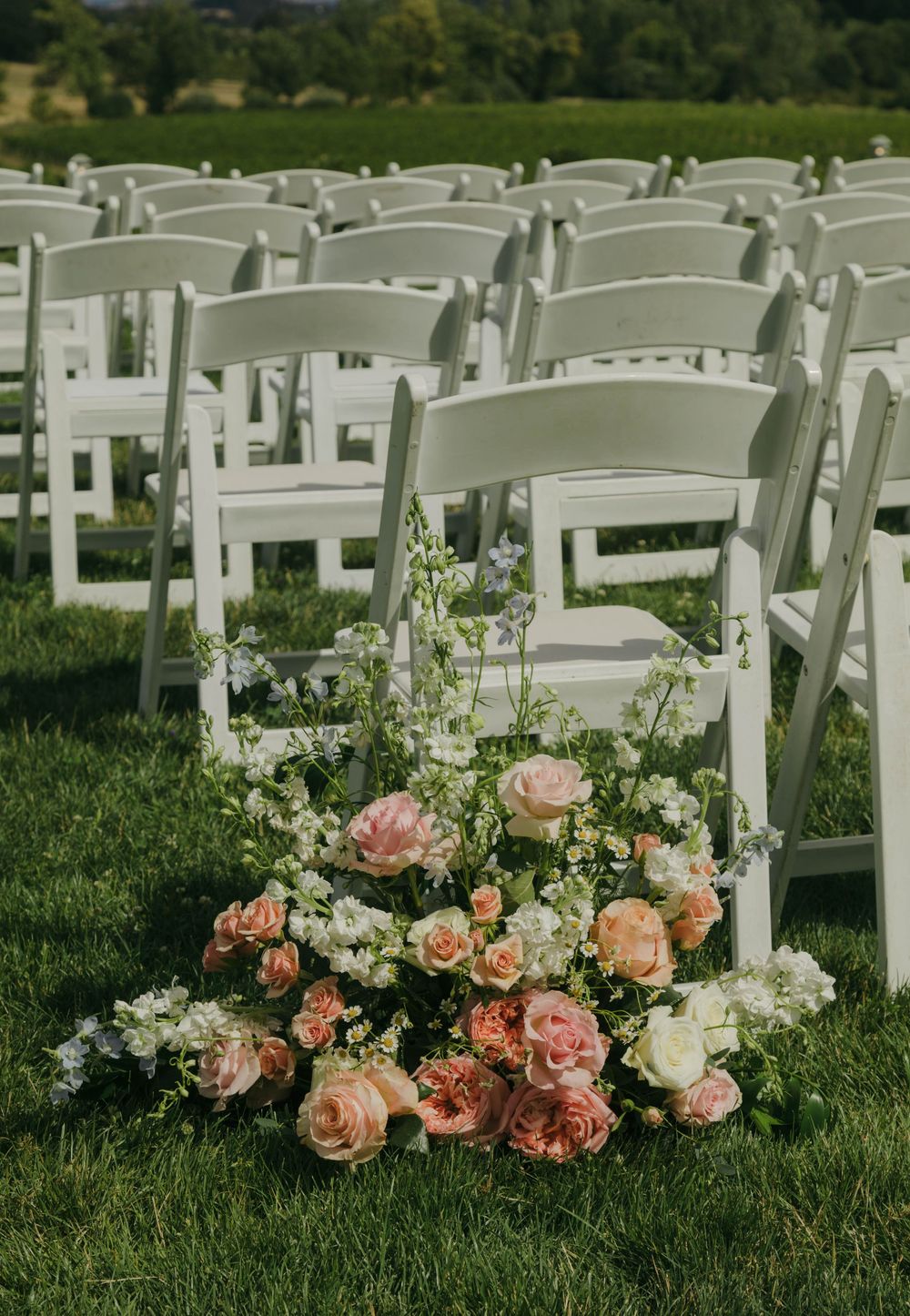 White folding chairs arranged on green grass with a lush floral arrangement of peach and pink roses at the aisle.